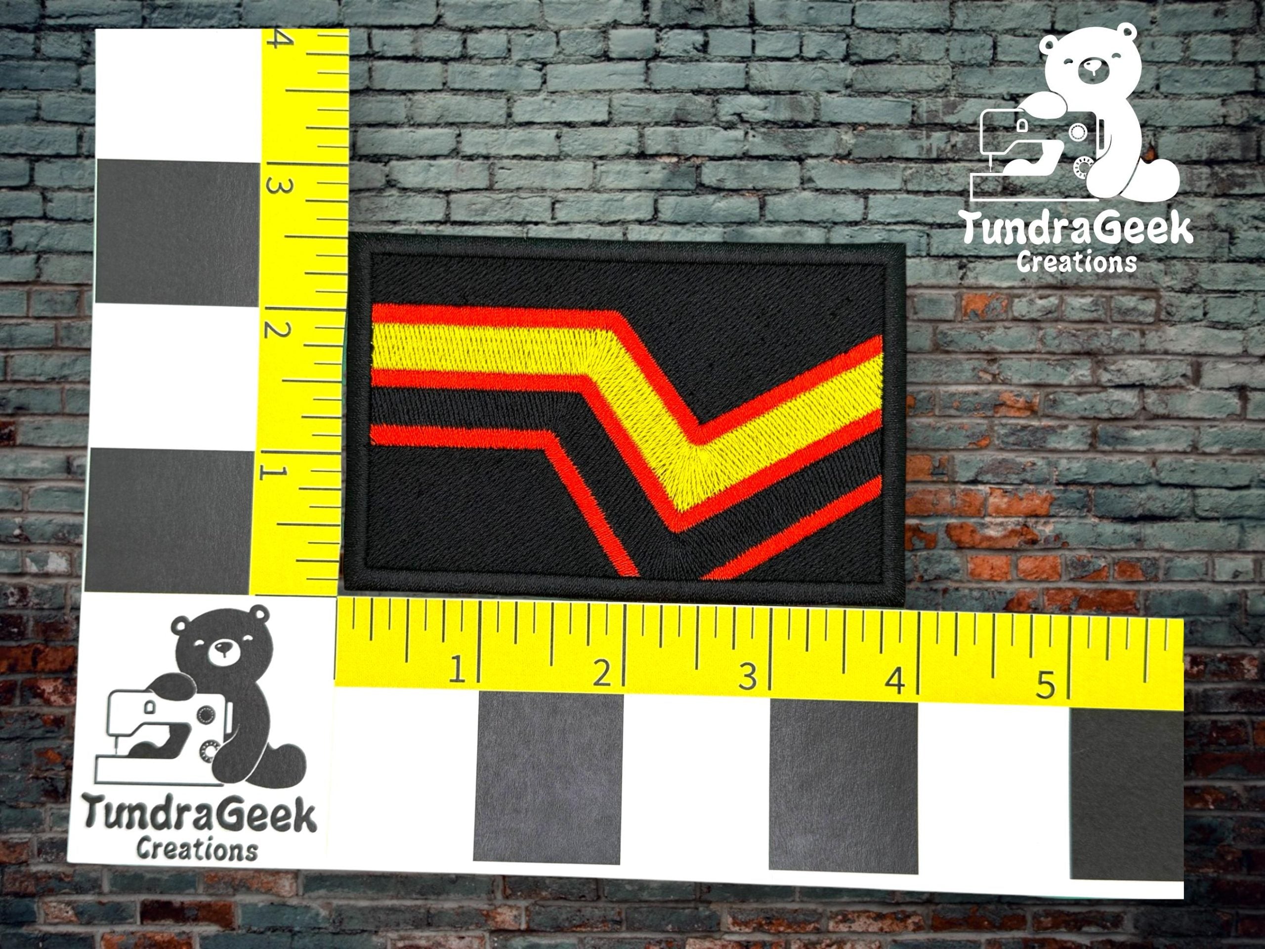 A rectangular embroidered patch with yellow and red chevron stripes, placed on a ruler grid for scale, displayed against a brick wall background.