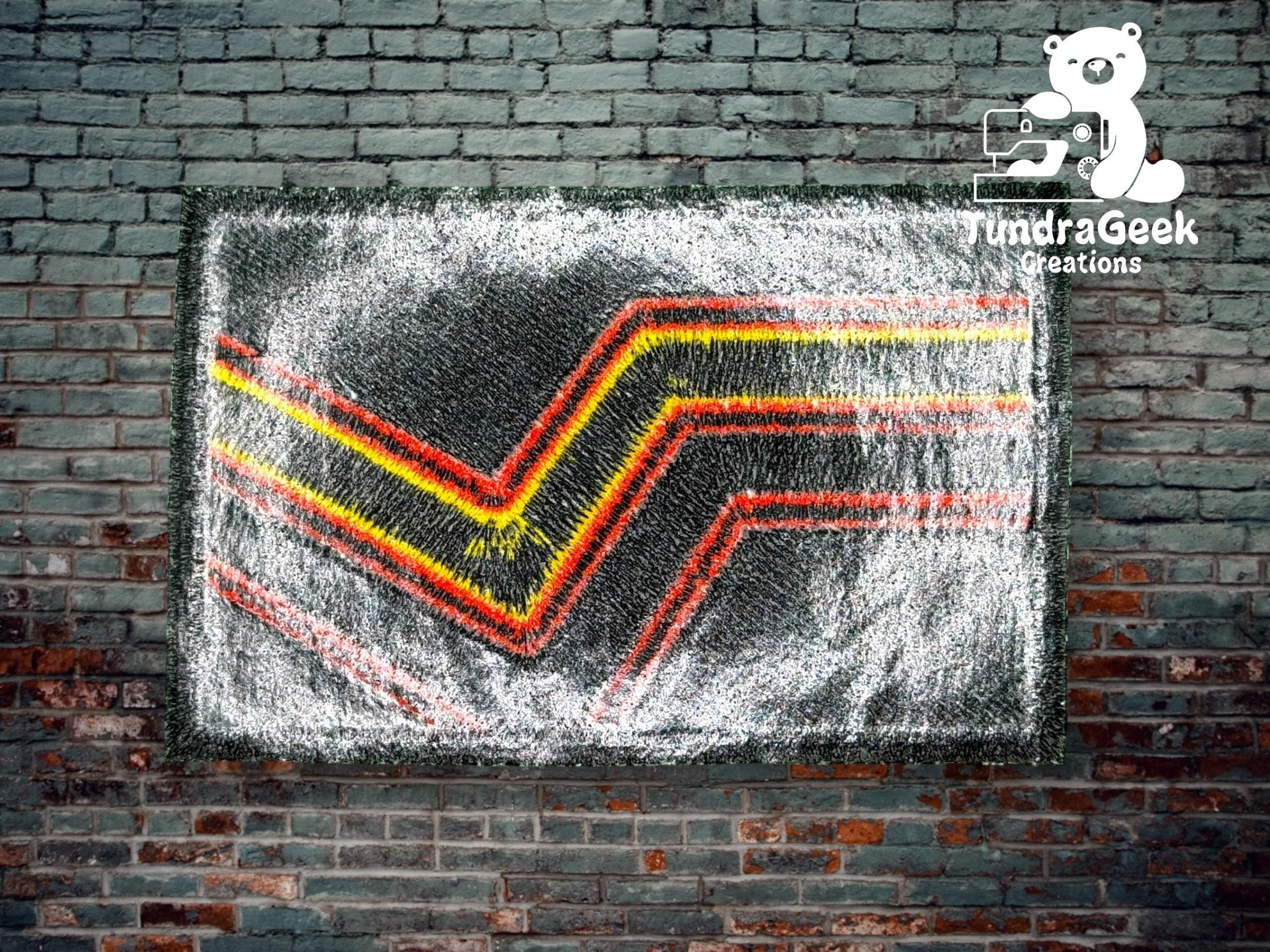 A piece of knit art with red, yellow, and black chevron stripes hangs on a brick wall, featuring a logo with a bear and sewing machine in the top right corner.