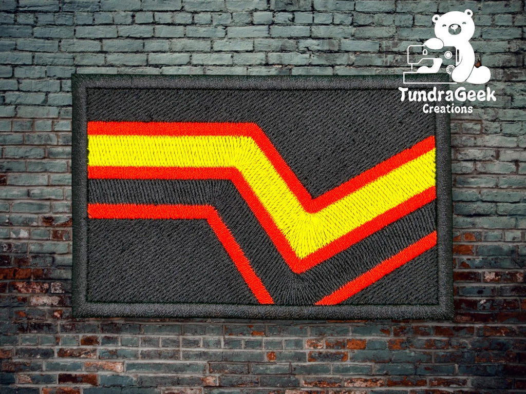 A rectangular patch with a zigzag red, yellow, and orange stripe on a black background, displayed against a brick wall with the TundraGeek Creations logo in the top right corner.