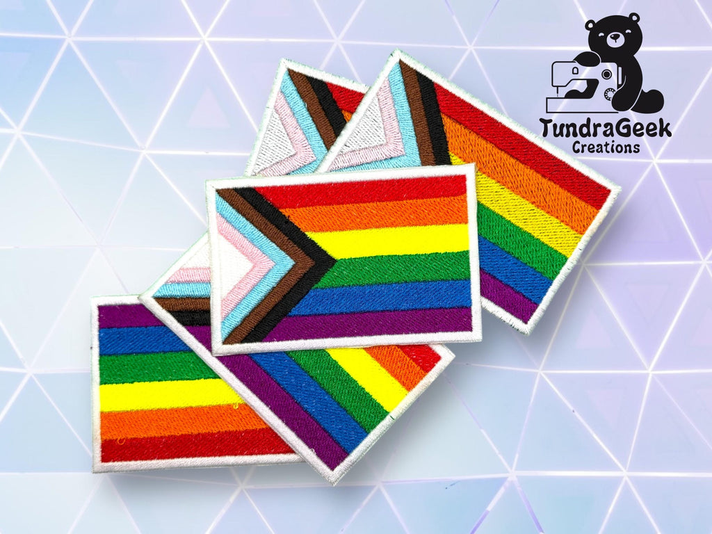 Embroidered progress pride flag patches overlap against a geometric pastel background, with the TundraGeek Creations logo featuring a bear and sewing machine in the top right corner.