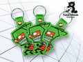 Three green keychains featuring a cartoon character with red eyes and a wide grin, placed on a geometric white background. Tundra Geek Creations logo is in the top right corner.