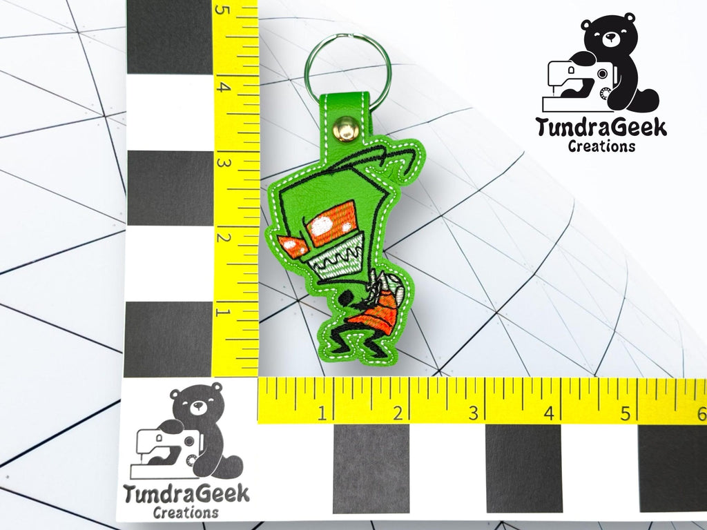 A green cartoon alien keychain with red eyes and a red shirt, displayed on a grid with ruler measurements in inches, next to a TundraGeek Creations logo.