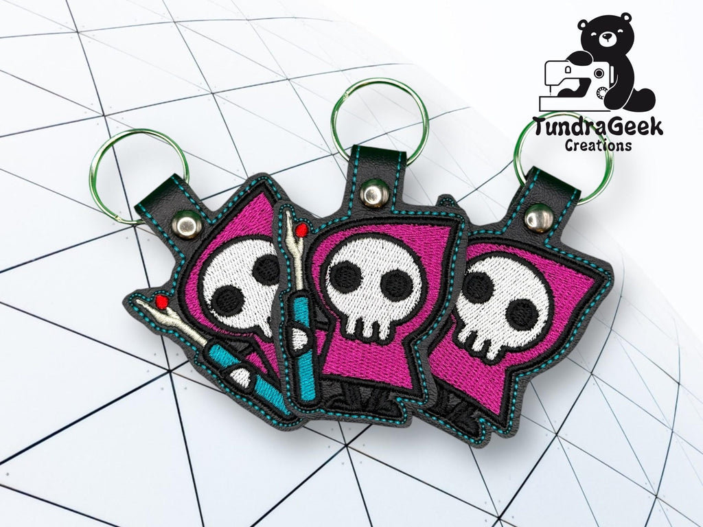 Three embroidered keychains featuring cartoon grim reapers in purple hooded cloaks, placed on a geometric white surface. TundraGeek Creations logo appears in the top right corner.
