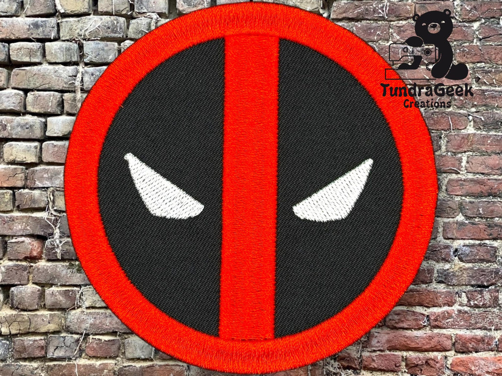 A circular patch with a red border and stripe, black background, and two white eye shapes, resembling the Deadpool logo, displayed against a brick wall background.
