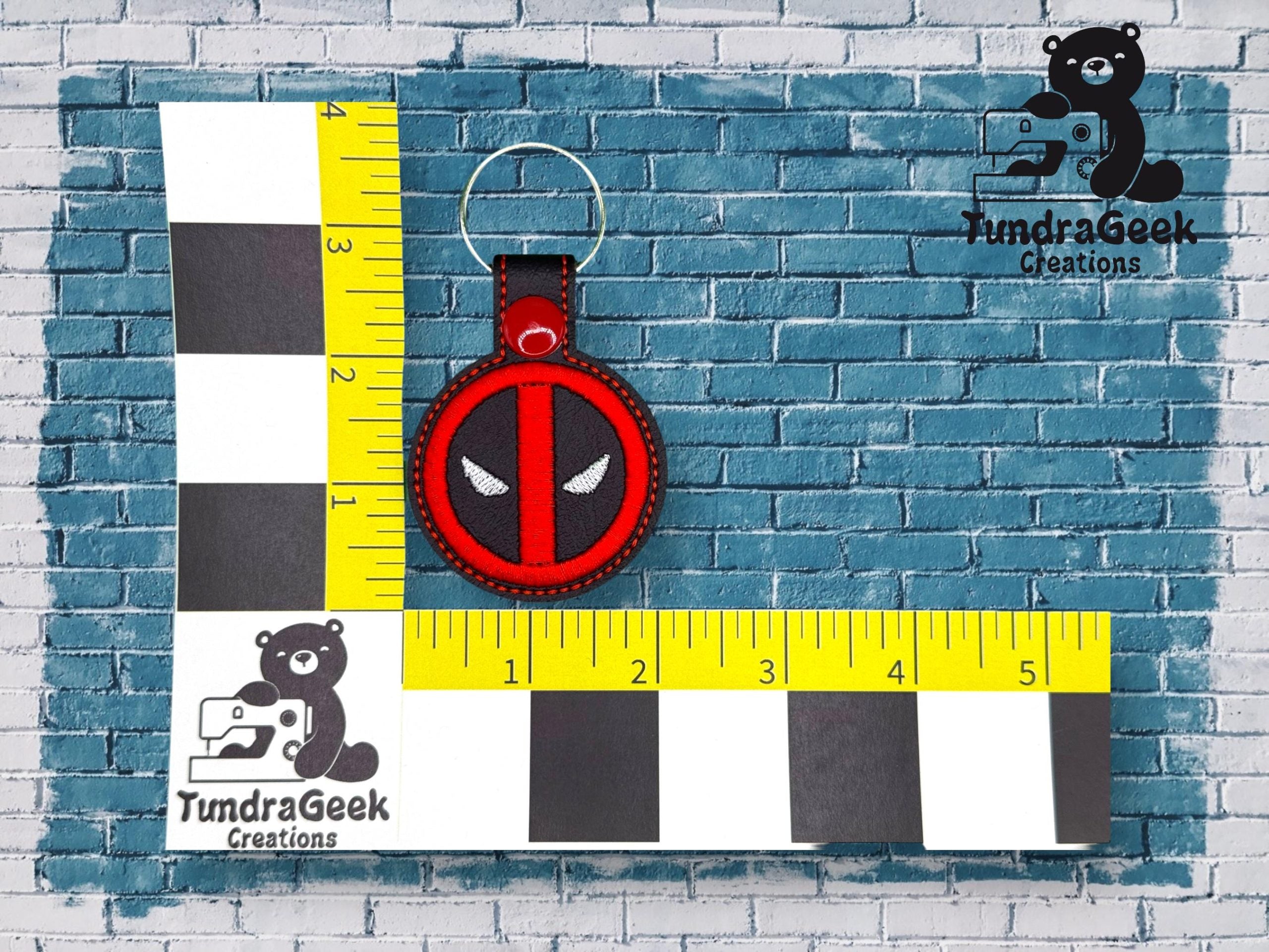 A round keychain with a red and black design resembling Deadpool’s mask is displayed on a measurement grid against a blue brick background.