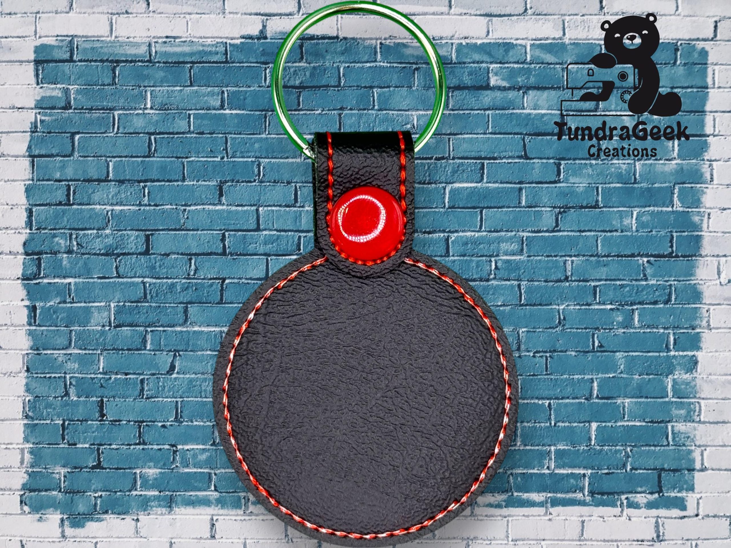 A round black keychain with red stitching and a green metal ring is displayed against a blue brick wall with the TundraGeek Creations logo in the top right corner.
