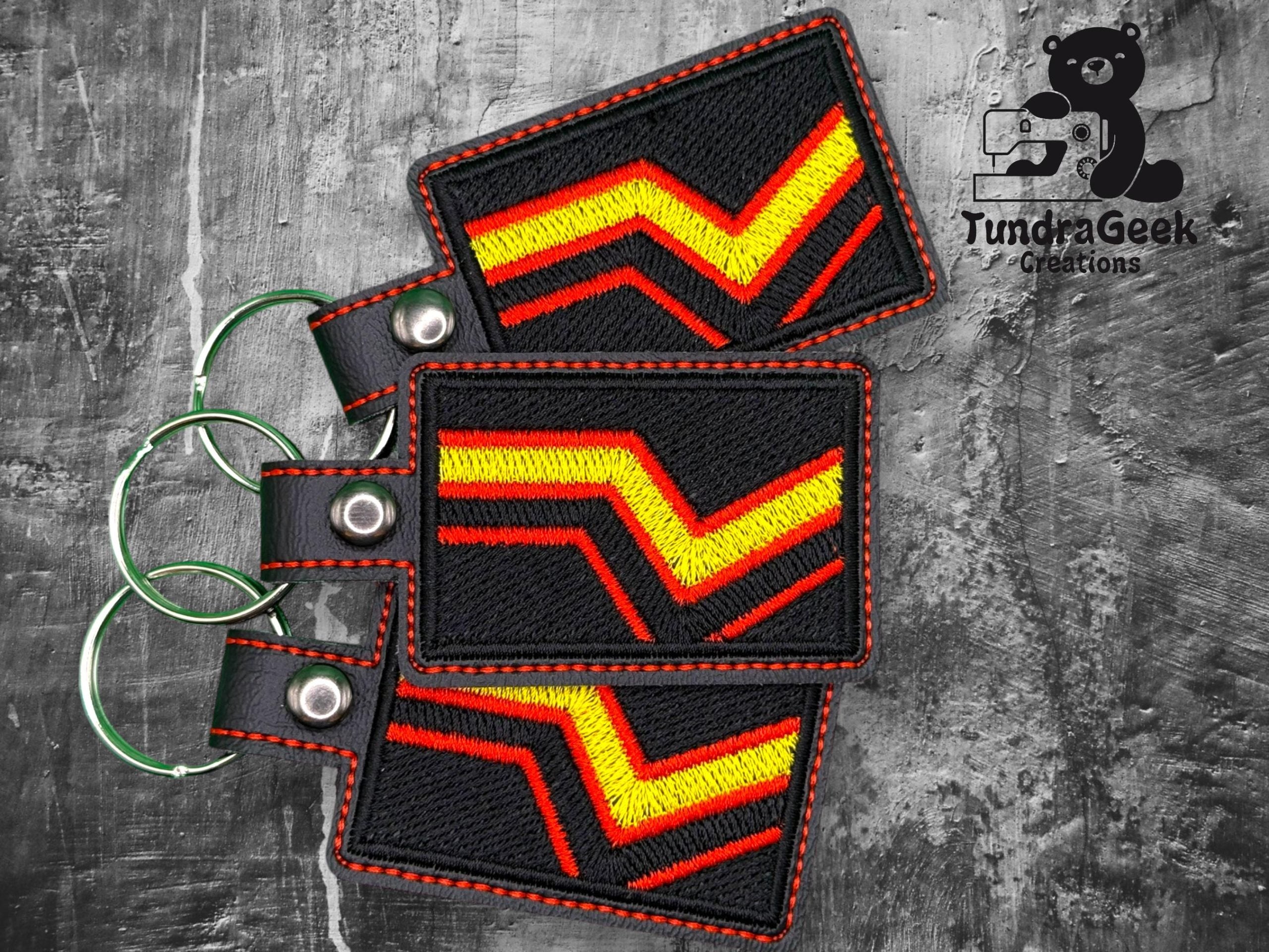 Three rectangular keychains with red, yellow, and orange chevron patterns, displayed on a gray textured surface. TundraGeek Creations logo is visible in the upper right corner.