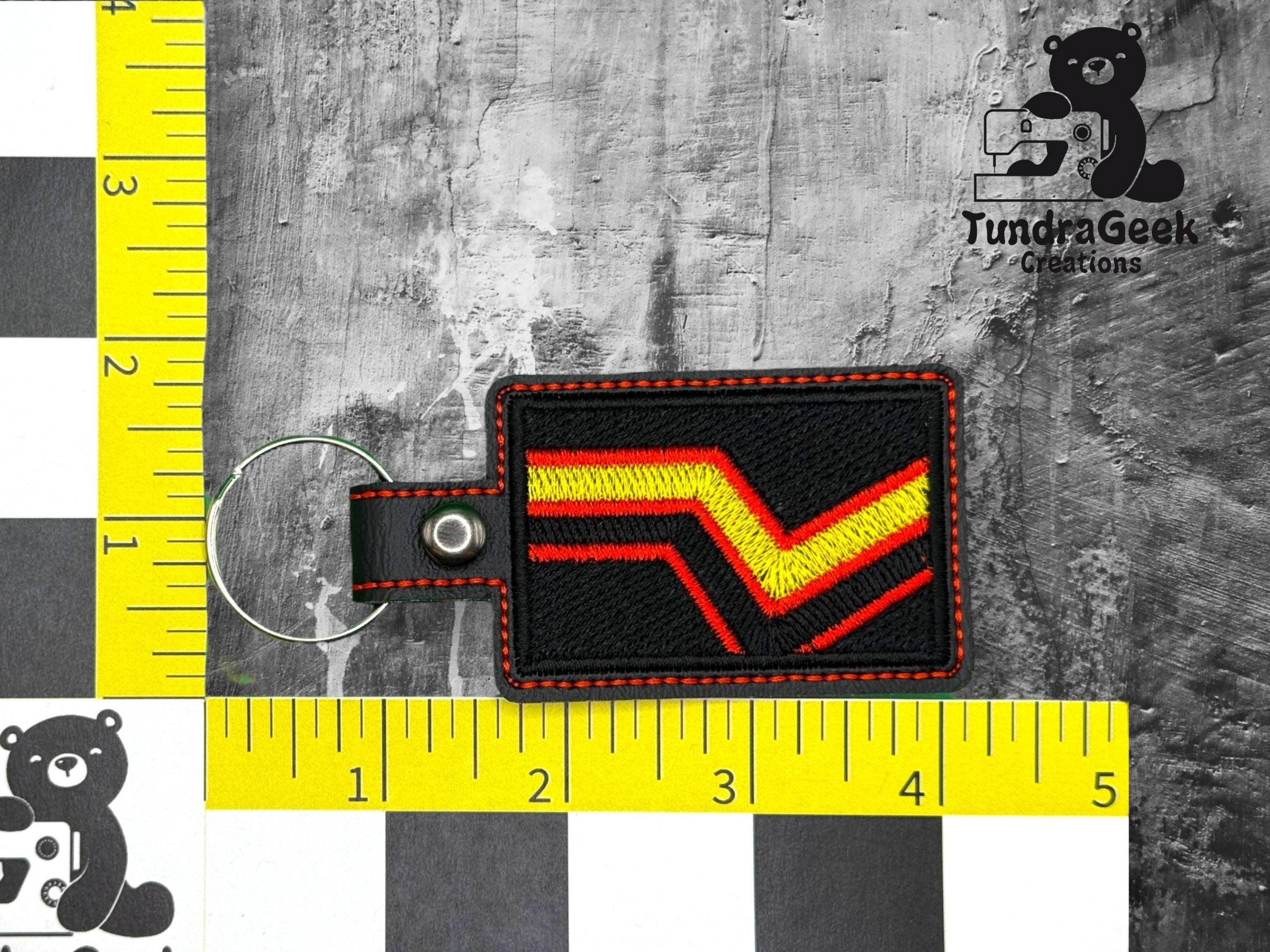 A rectangular keychain with red and yellow chevron embroidery, black background, and red stitching, placed on a ruler grid for scale. TundraGeek Creations logo in the top right corner.