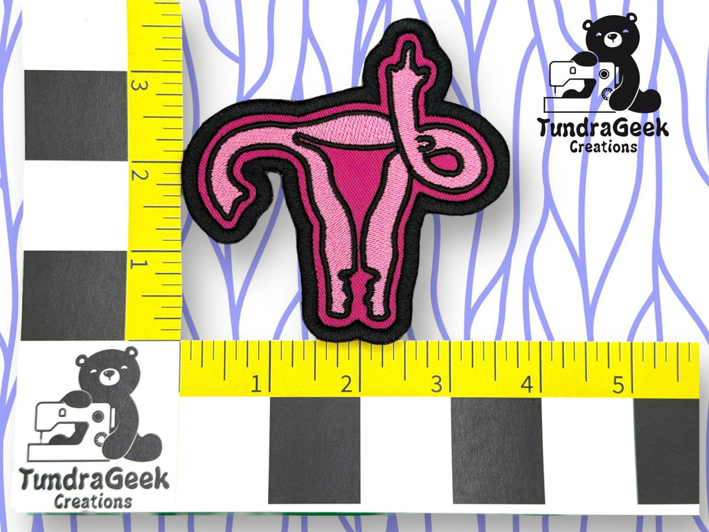 An embroidered patch of a pink uterus is placed between yellow rulers for scale. The TundraGeek Creations logo featuring a bear with a sewing machine is visible in two corners.