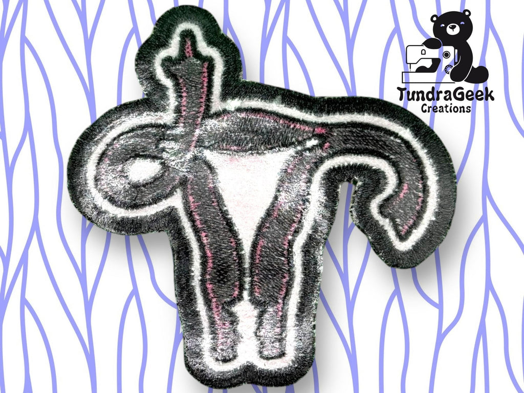An embroidered uterus patch with one fallopian tube shaped as a raised middle finger, shown on a blue patterned background with the TundraGeek Creations logo.