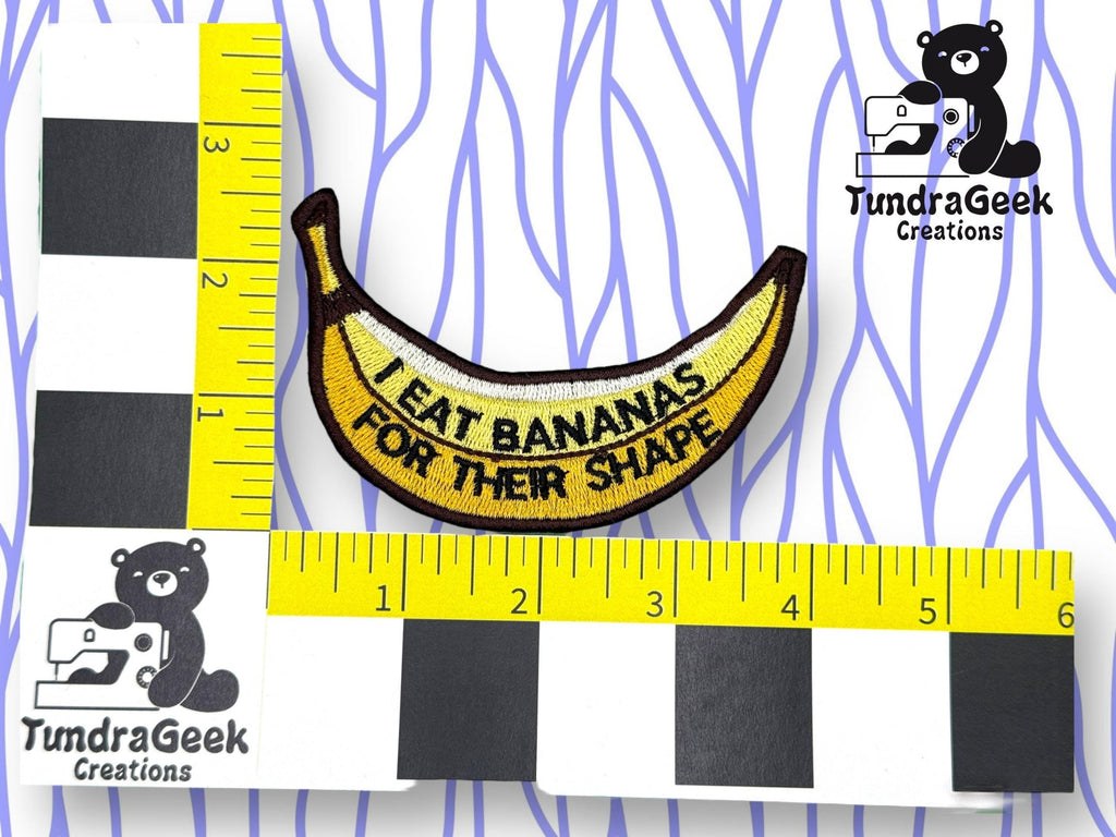 A banana-shaped patch with the text I eat bananas for their shape is placed between two yellow rulers on a white surface with a blue wavy line pattern and TundraGeek Creations logos.