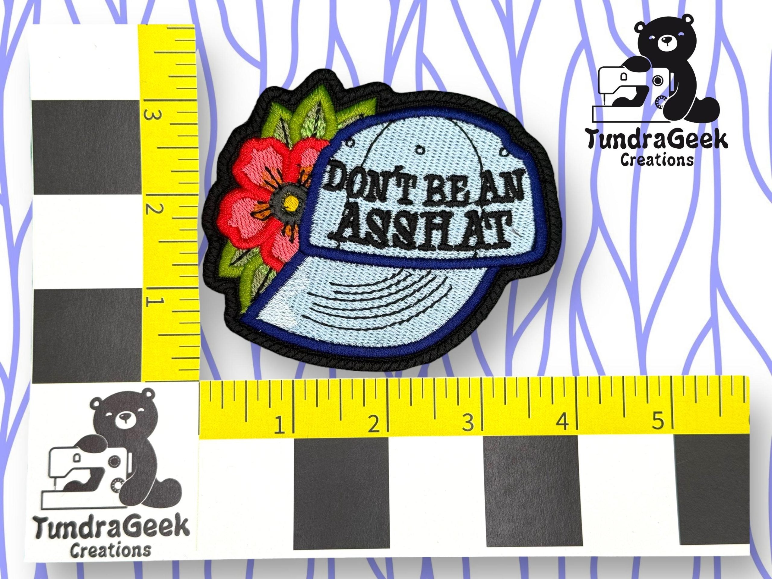 An embroidered patch of a blue cap with a red flower and the words DONT BE AN ASSHAT, next to rulers for scale. TundraGeek Creations logo appears in two corners.