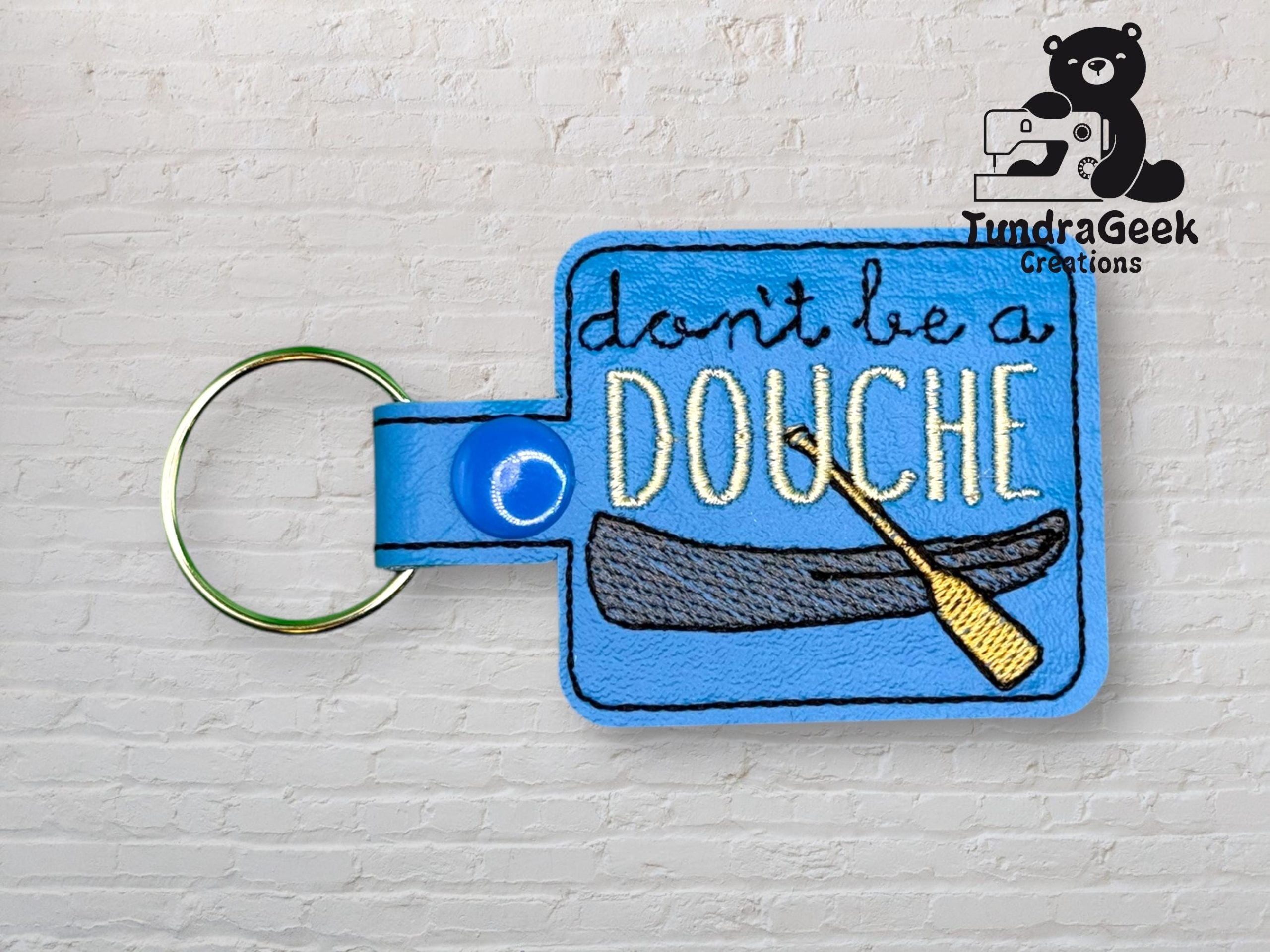 A blue keychain with embroidered text dont be a DOUCHE above an image of a canoe and paddle. TundraGeek Creations logo appears in the top right corner.