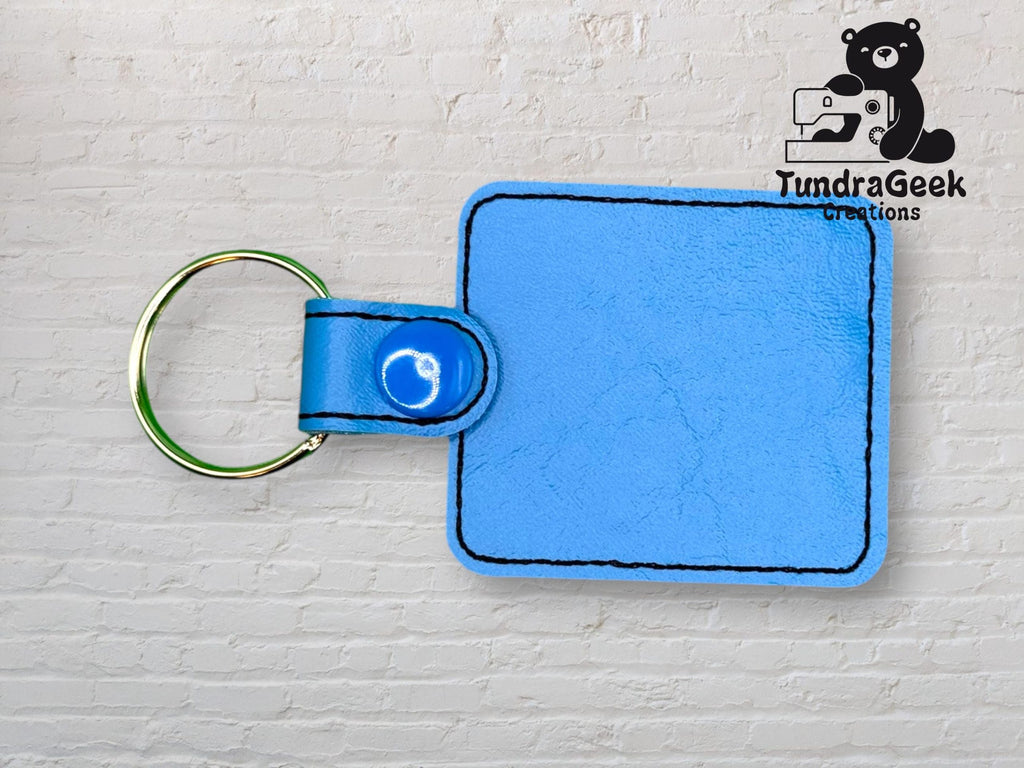 A square light blue faux leather keychain with black stitching and a gold keyring, displayed against a white brick background. The TundraGeek Creations logo is in the upper right corner.