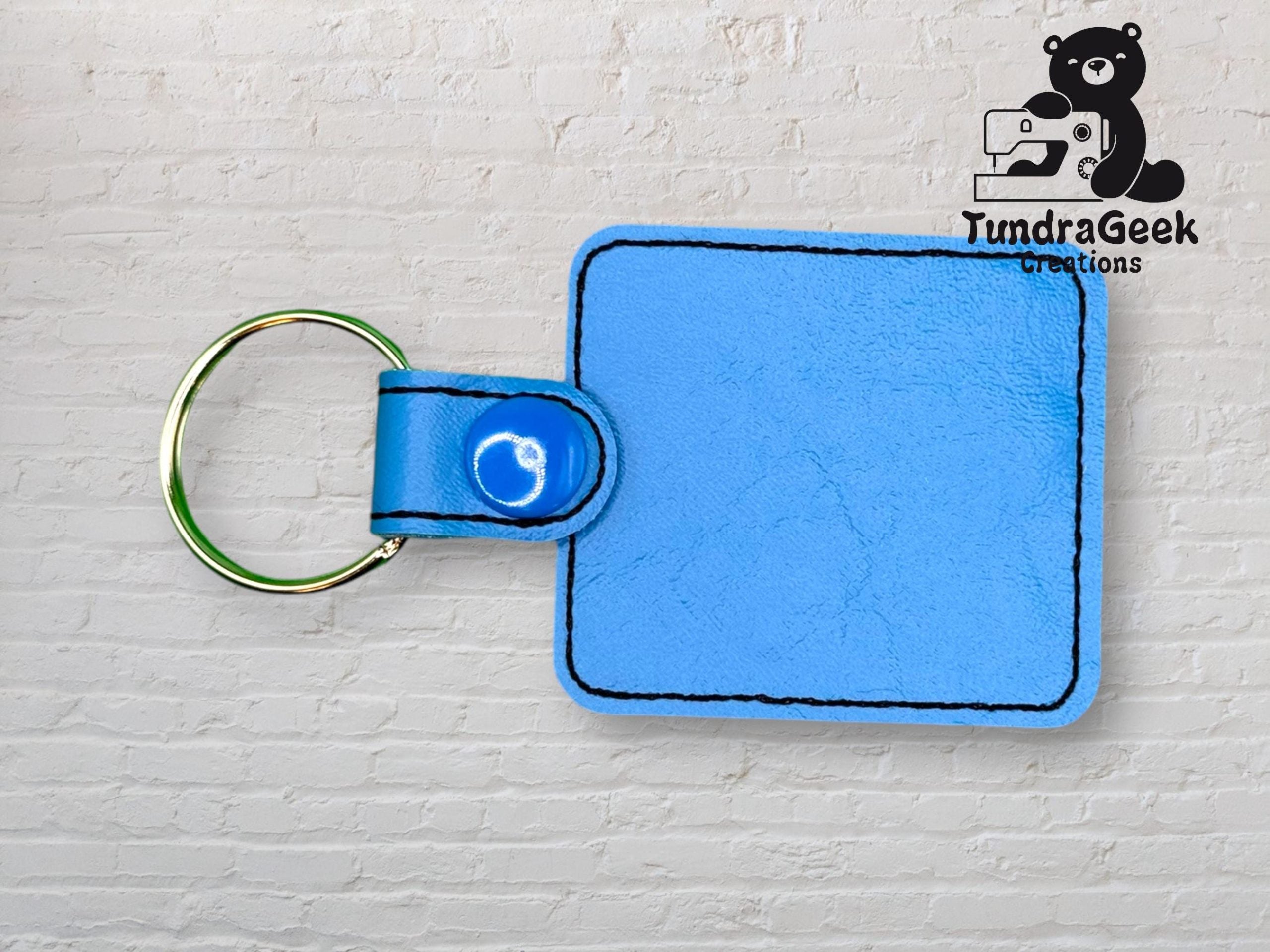 A square light blue faux leather keychain with black stitching and a gold keyring, displayed against a white brick background. The TundraGeek Creations logo is in the upper right corner.