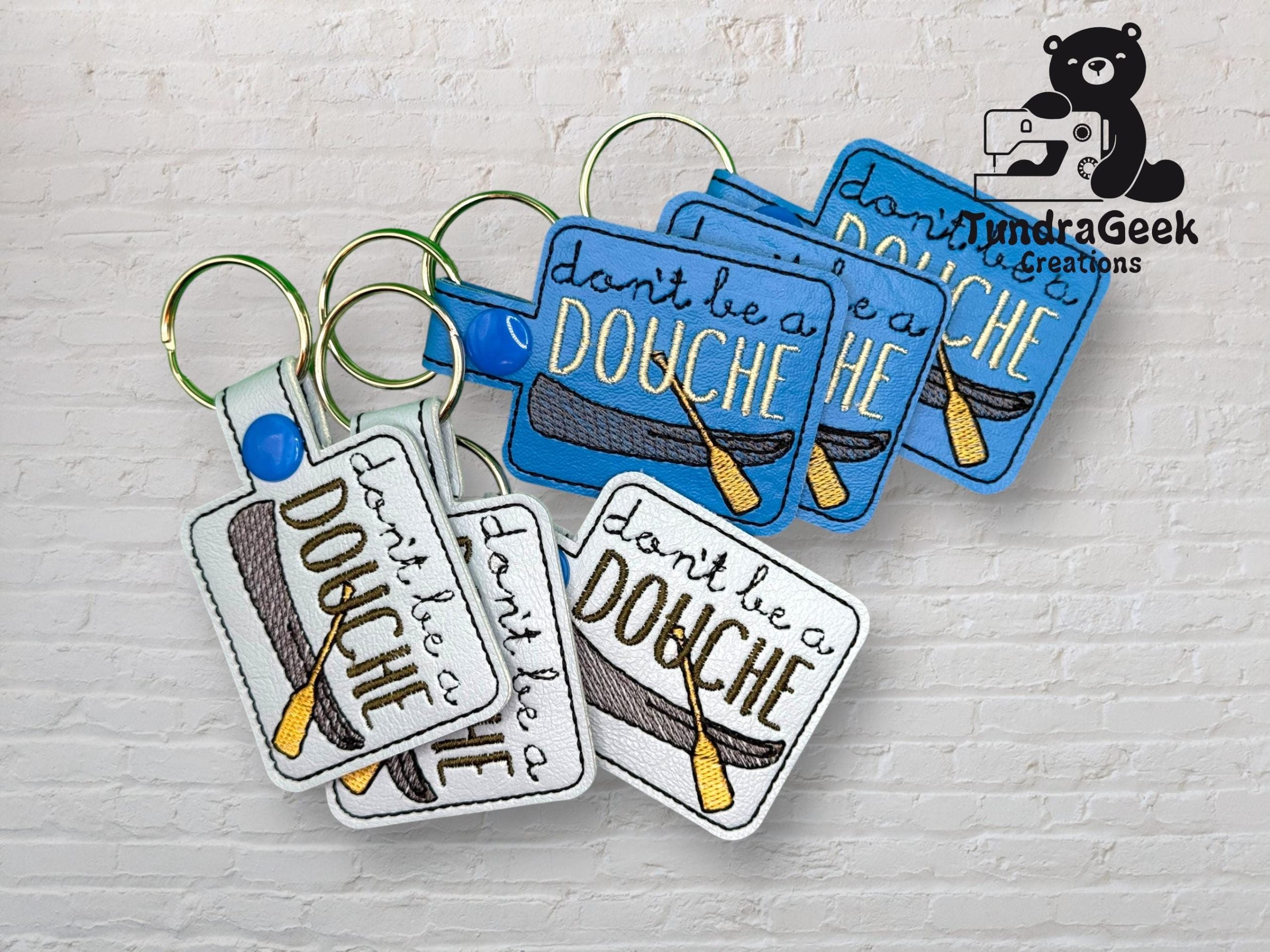 Four keychains with embroidered text reading dont be a DOUCHE and an illustration of a douche, displayed against a white brick wall with the TundraGeek Creations logo in the corner.