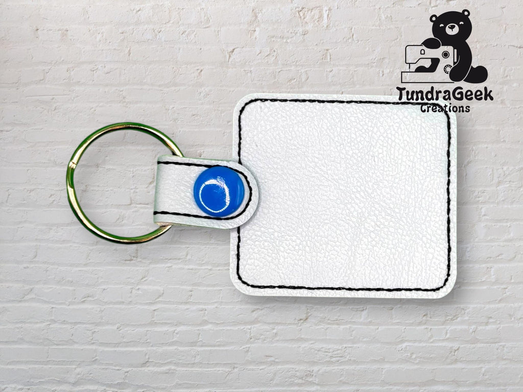 White square leather keychain with black stitching and blue snap, placed on a light brick background. TundraGeek Creations logo with bear and sewing machine in the top right corner.