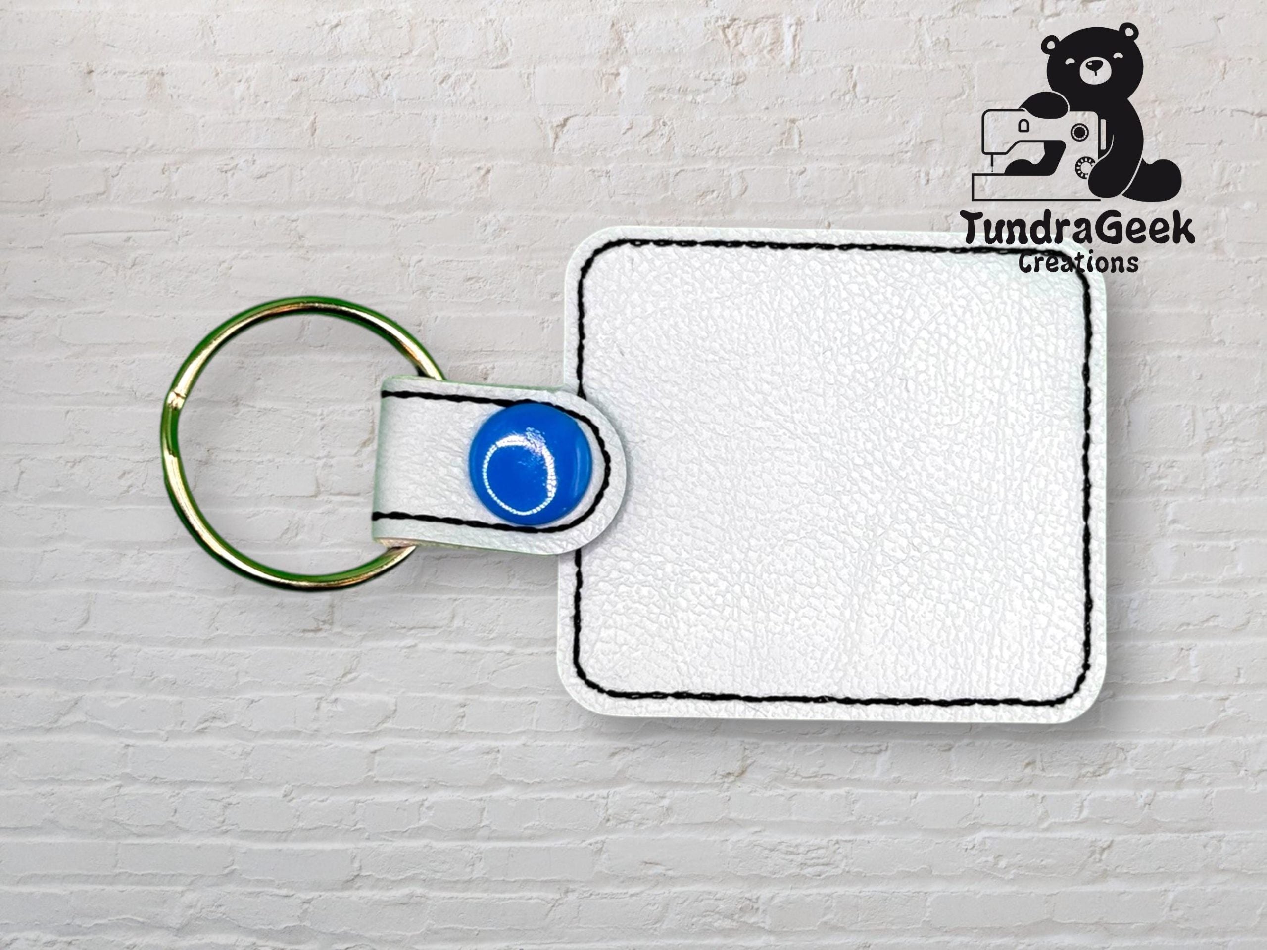 White square leather keychain with black stitching and blue snap, placed on a light brick background. TundraGeek Creations logo with bear and sewing machine in the top right corner.