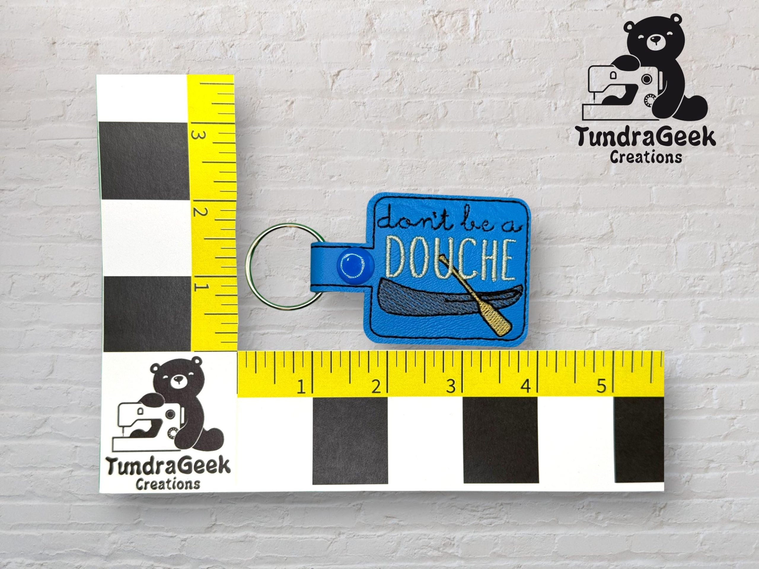 A blue keychain with text dont be a DOUCHE and a canoe graphic, displayed next to a yellow ruler for scale, on a white brick background.