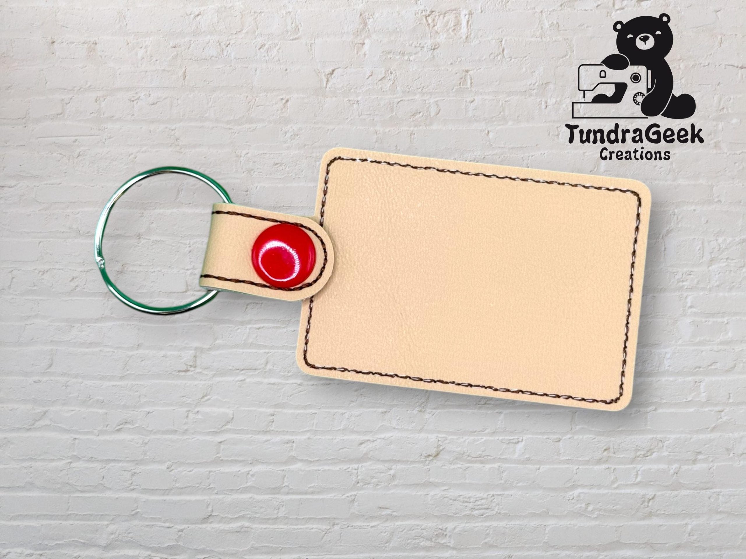 Rectangular beige leather keychain with a metal ring and red snap, placed on a white brick background. TundraGeek Creations logo with a bear and sewing machine is in the top right corner.