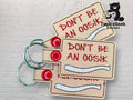 Three beige keychains with metal rings and red snaps display the text DONT BE AN OOSH in red, with a white embroidered bone shape below the text.