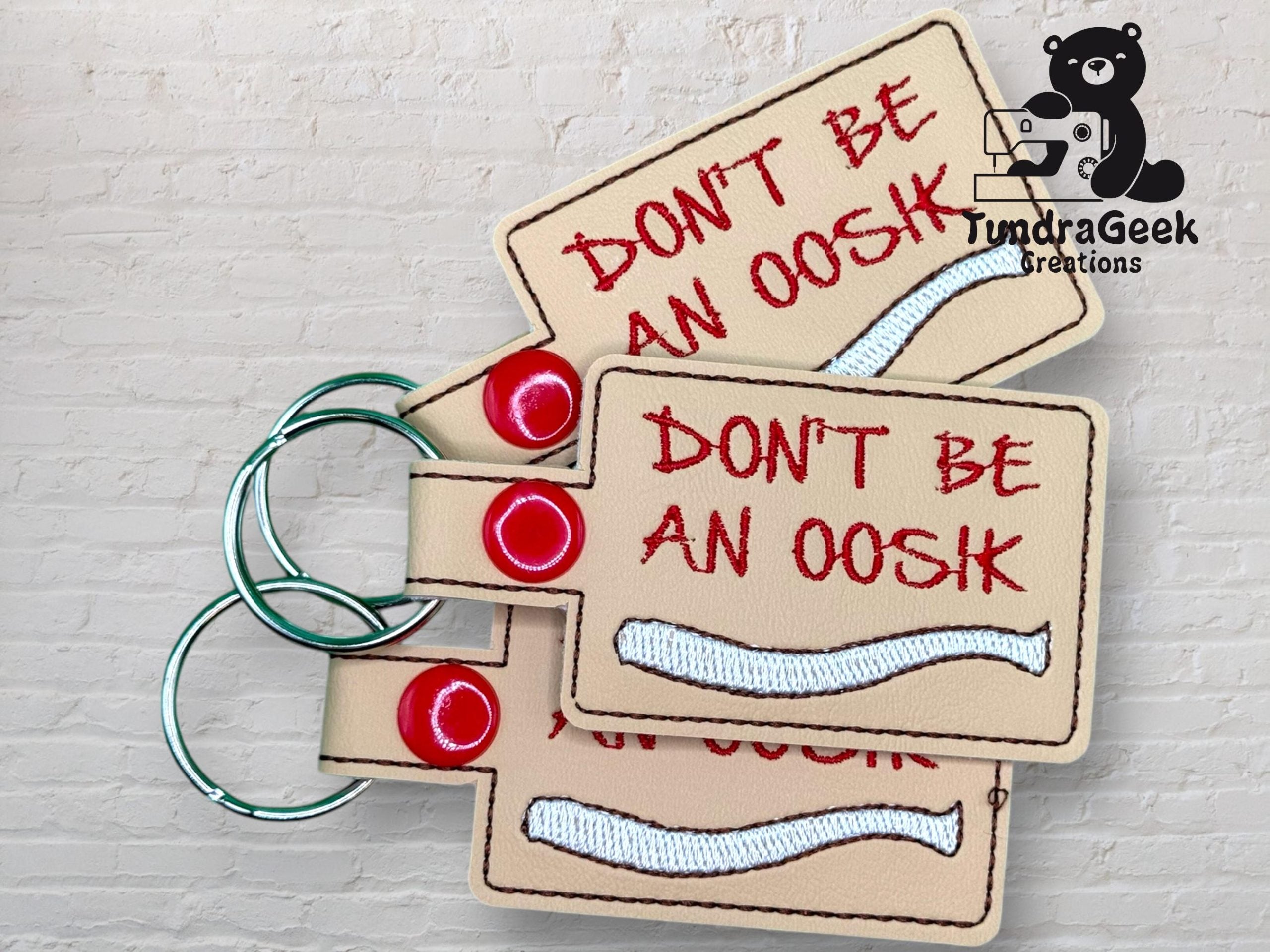 Three beige keychains with metal rings and red snaps display the text DONT BE AN OOSH in red, with a white embroidered bone shape below the text.