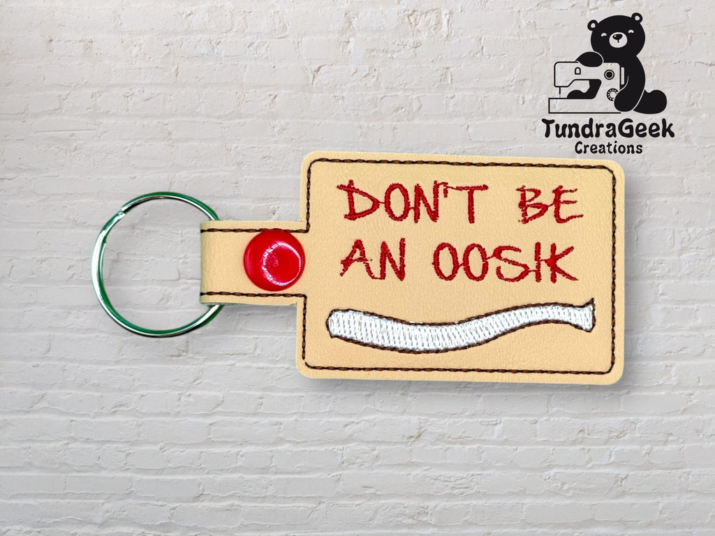 A beige keychain with DONT BE AN OOSHK embroidered in red, a white bone design, and a red snap button, placed on a light brick background with the TundraGeek Creations logo.