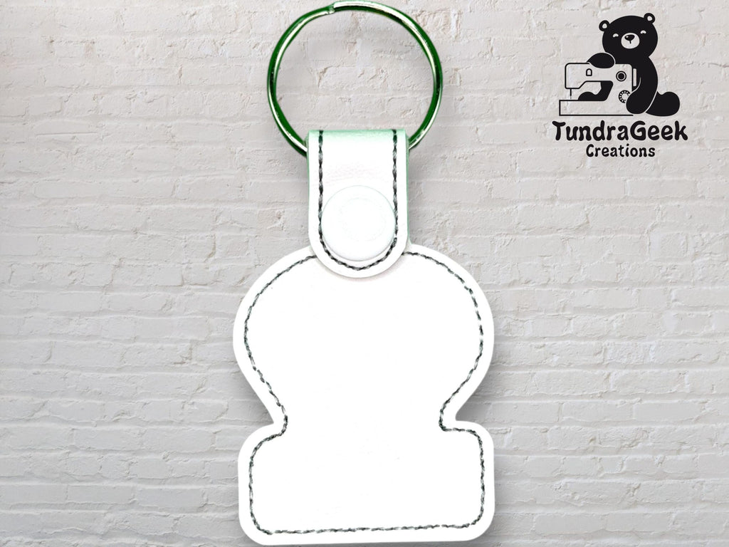 White blank keychain with black stitching and a green ring, displayed in front of a white brick wall with the TundraGeek Creations logo in the top right corner.