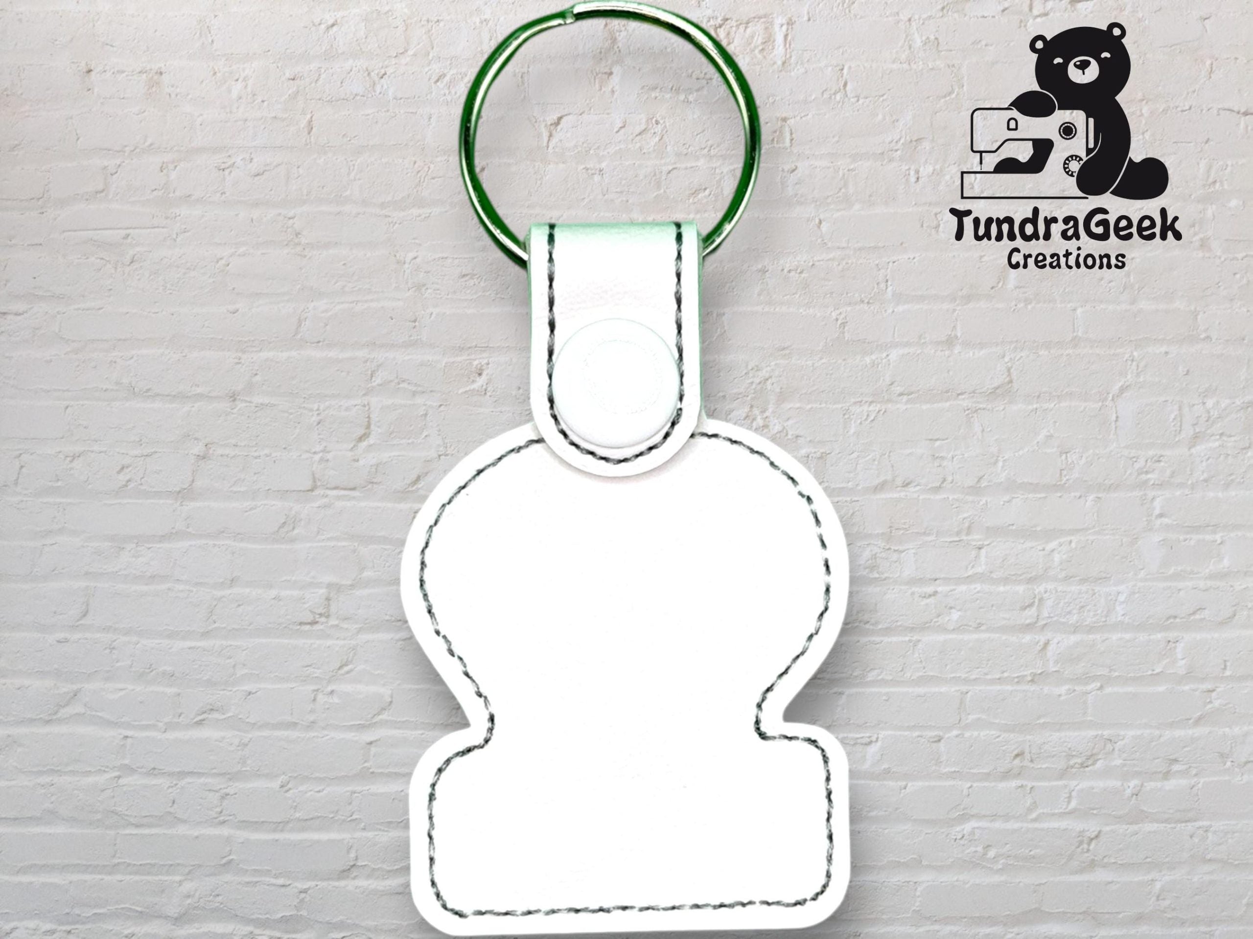 White blank keychain with black stitching and a green ring, displayed in front of a white brick wall with the TundraGeek Creations logo in the top right corner.