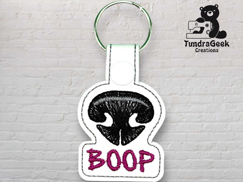 A white keychain with black embroidery of a bear nose above the word BOOP in purple, displayed against a white brick wall with a TundraGeek Creations logo.