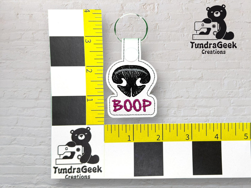 Keychain featuring an embroidered black animal nose and the word BOOP in purple, shown against a ruler for scale. TundraGeek Creations logo appears in the image.