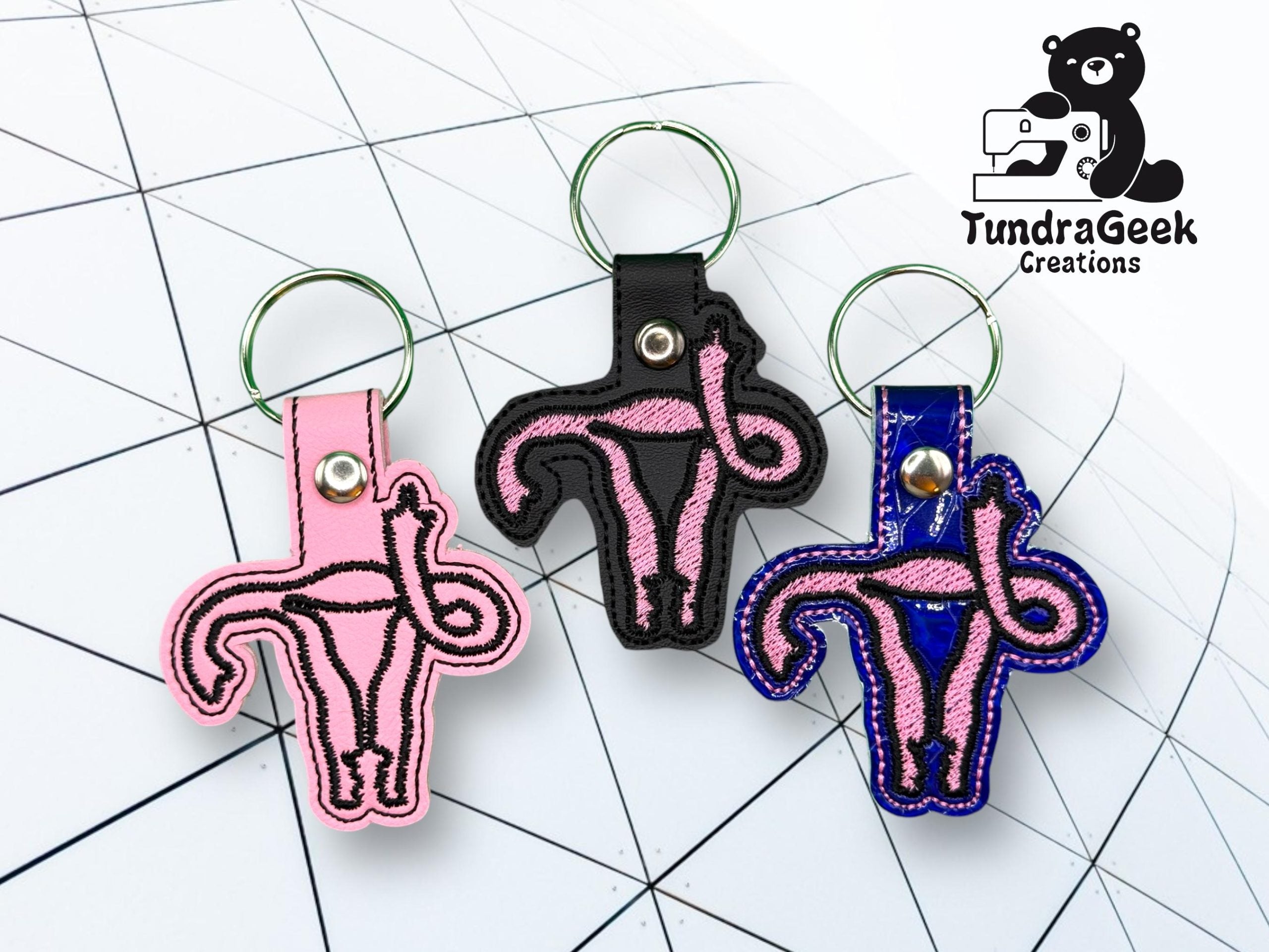 Three keychains featuring embroidered pink uterus designs on different colored backings are displayed on a geometric white surface. The TundraGeek Creations logo appears in the top right corner.