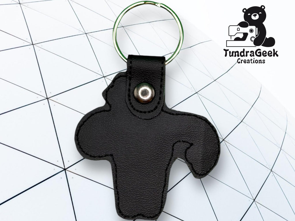 A black leather keychain shaped like a stylized animal, with a metal ring and snap, is placed on a geometric white background with the TundraGeek Creations logo in the corner.