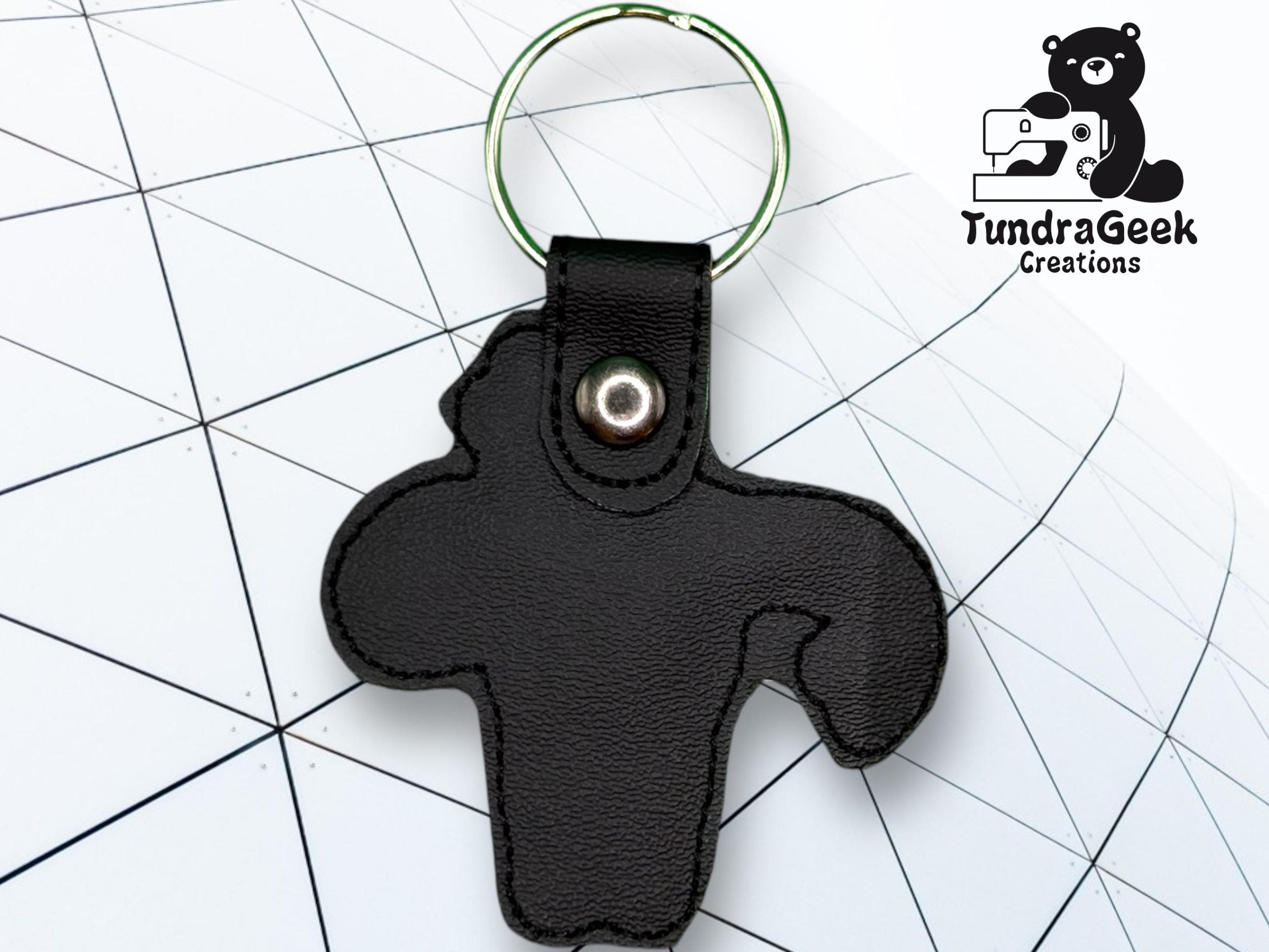 A black leather keychain shaped like a stylized animal, with a metal ring and snap, is placed on a geometric white background with the TundraGeek Creations logo in the corner.