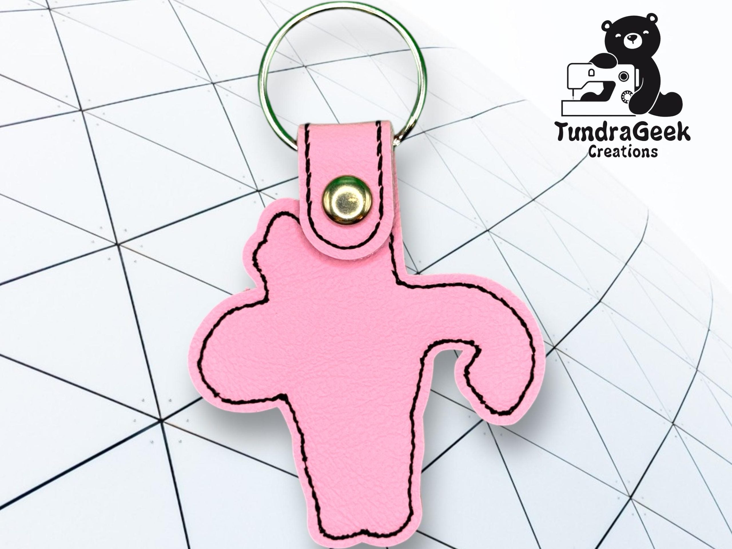 A pink keychain shaped like a uterus lies on a geometric-patterned surface. The logo TundraGeek Creations with a bear and sewing machine is in the top right corner.