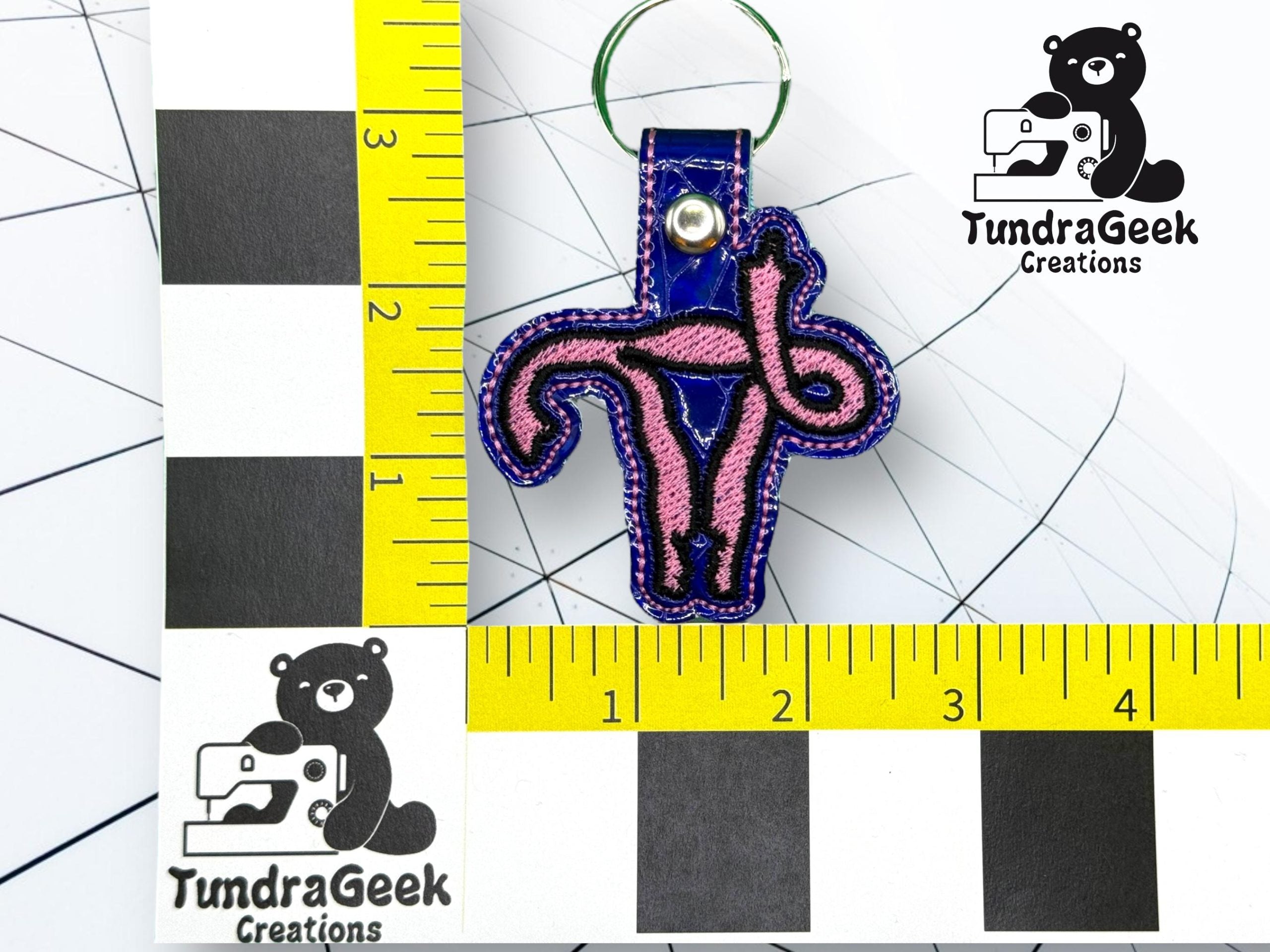 A keychain shaped like a pink and purple uterus is placed on a black and white grid with yellow rulers, featuring the TundraGeek Creations logo in the background.