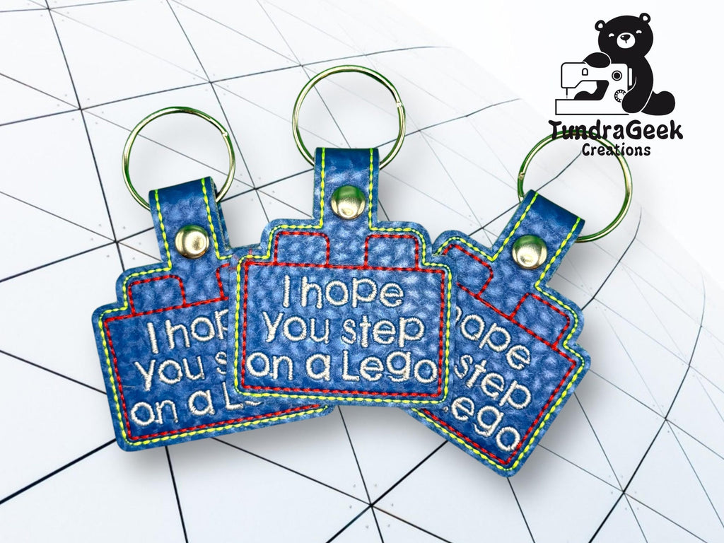 Three blue keychains with rainbow stitching read I hope you step on a Lego. They are arranged on a grid-patterned background, next to a TundraGeek Creations logo.