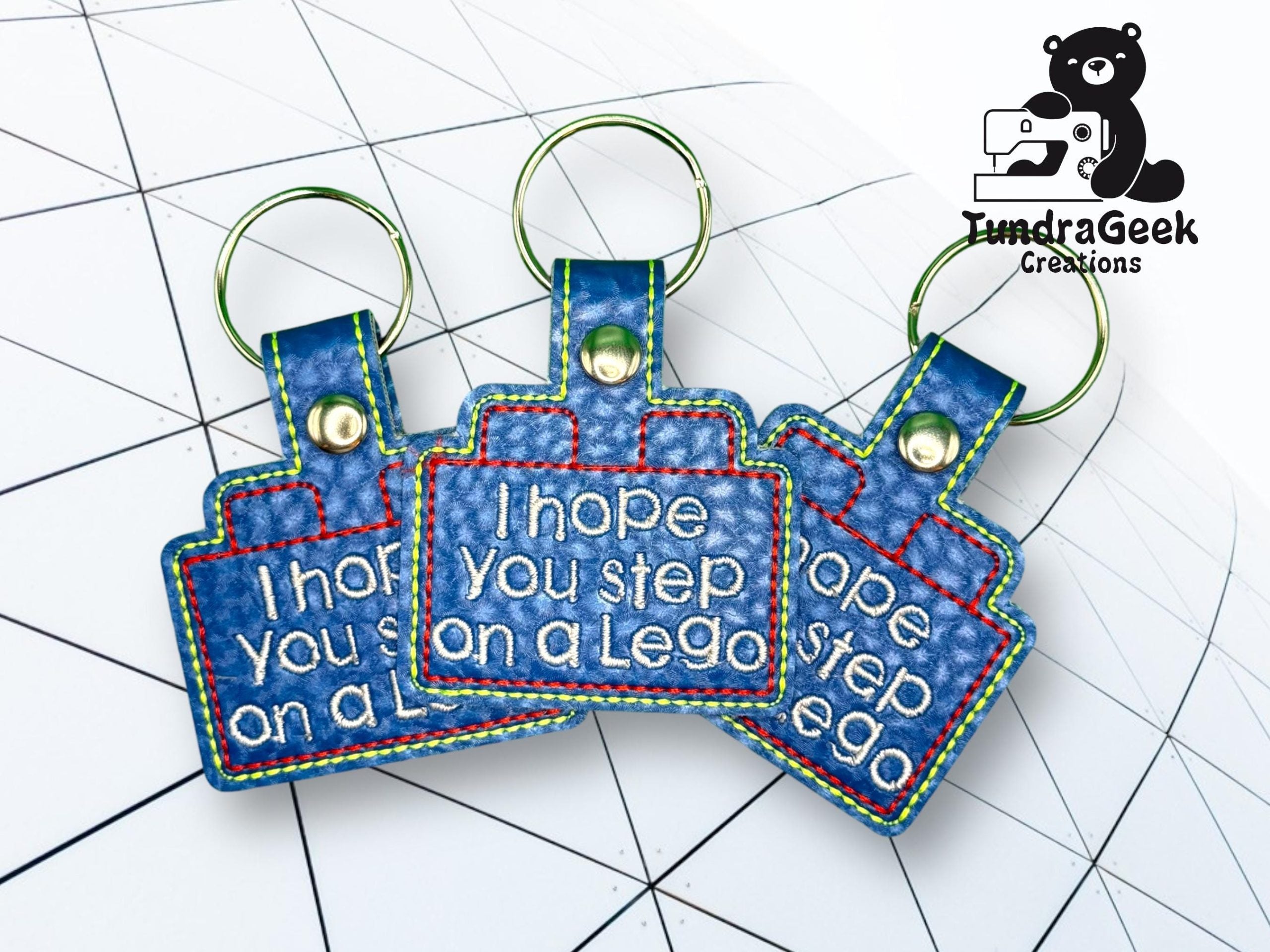 Three blue keychains with rainbow stitching read I hope you step on a Lego. They are arranged on a grid-patterned background, next to a TundraGeek Creations logo.