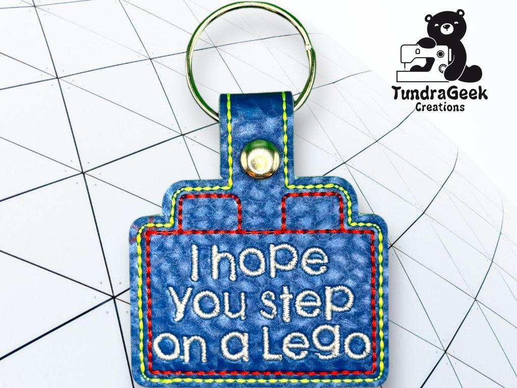 A blue keychain shaped like a Lego brick displays the embroidered text I hope you step on a Lego. A TundraGeek Creations logo is in the top right corner.