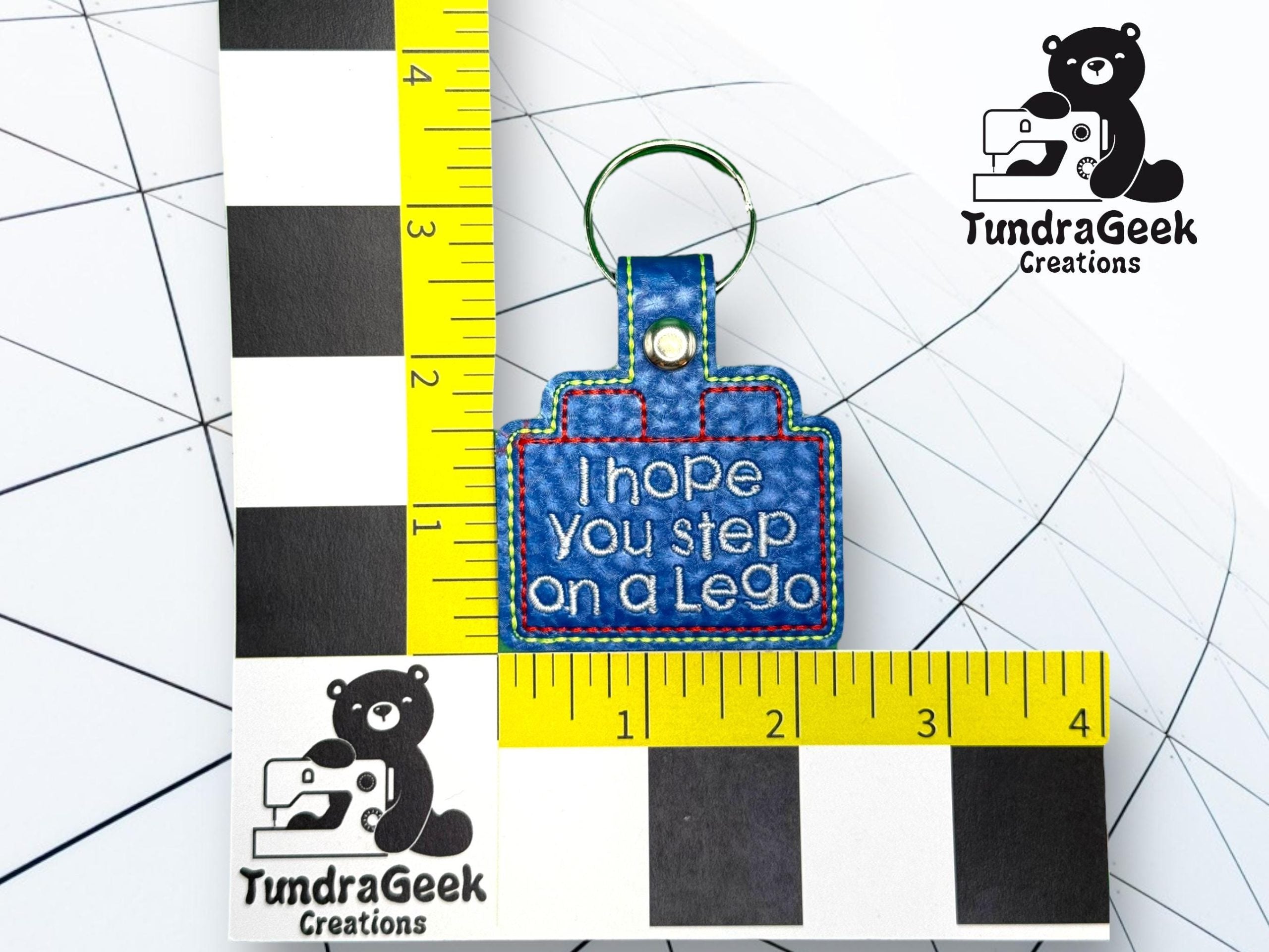 A blue keychain shaped like a LEGO brick reads I hope you step on a Lego. It is displayed on a measurement grid with TundraGeek Creations logos.