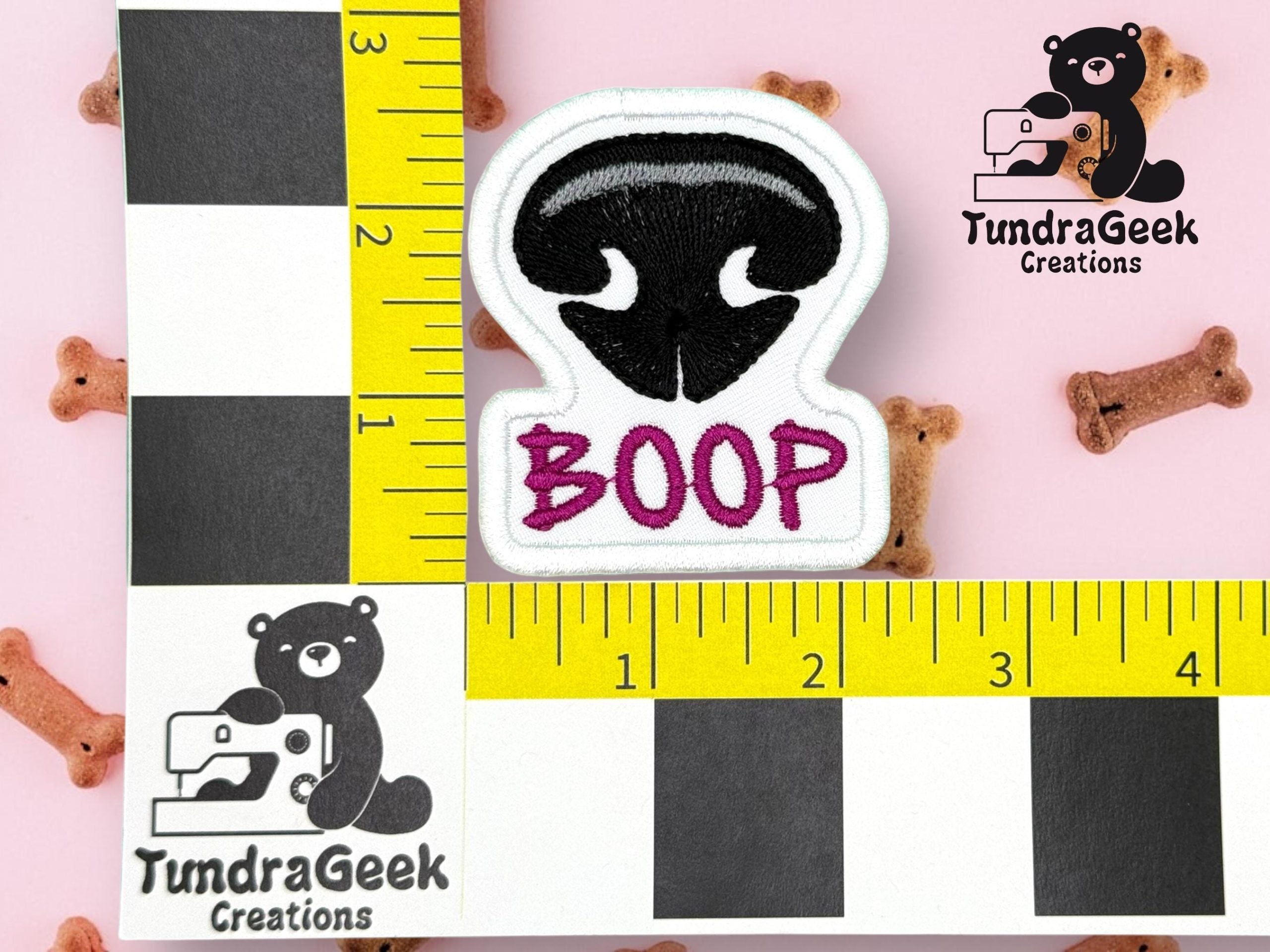 An embroidered patch shaped like a black dog nose with the word BOOP in pink, displayed on a pink background with small dog bone treats and measuring rulers.