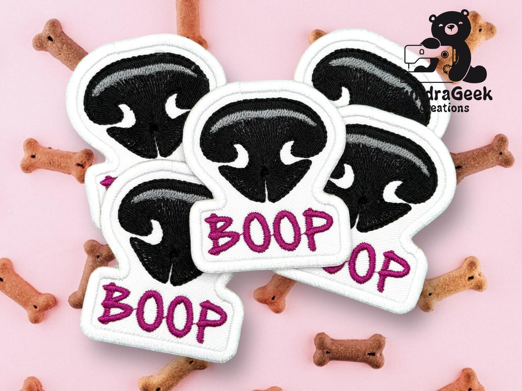 Five embroidered patches with a black animal nose and the word BOOP in pink, placed on a pink background with dog bone treats.