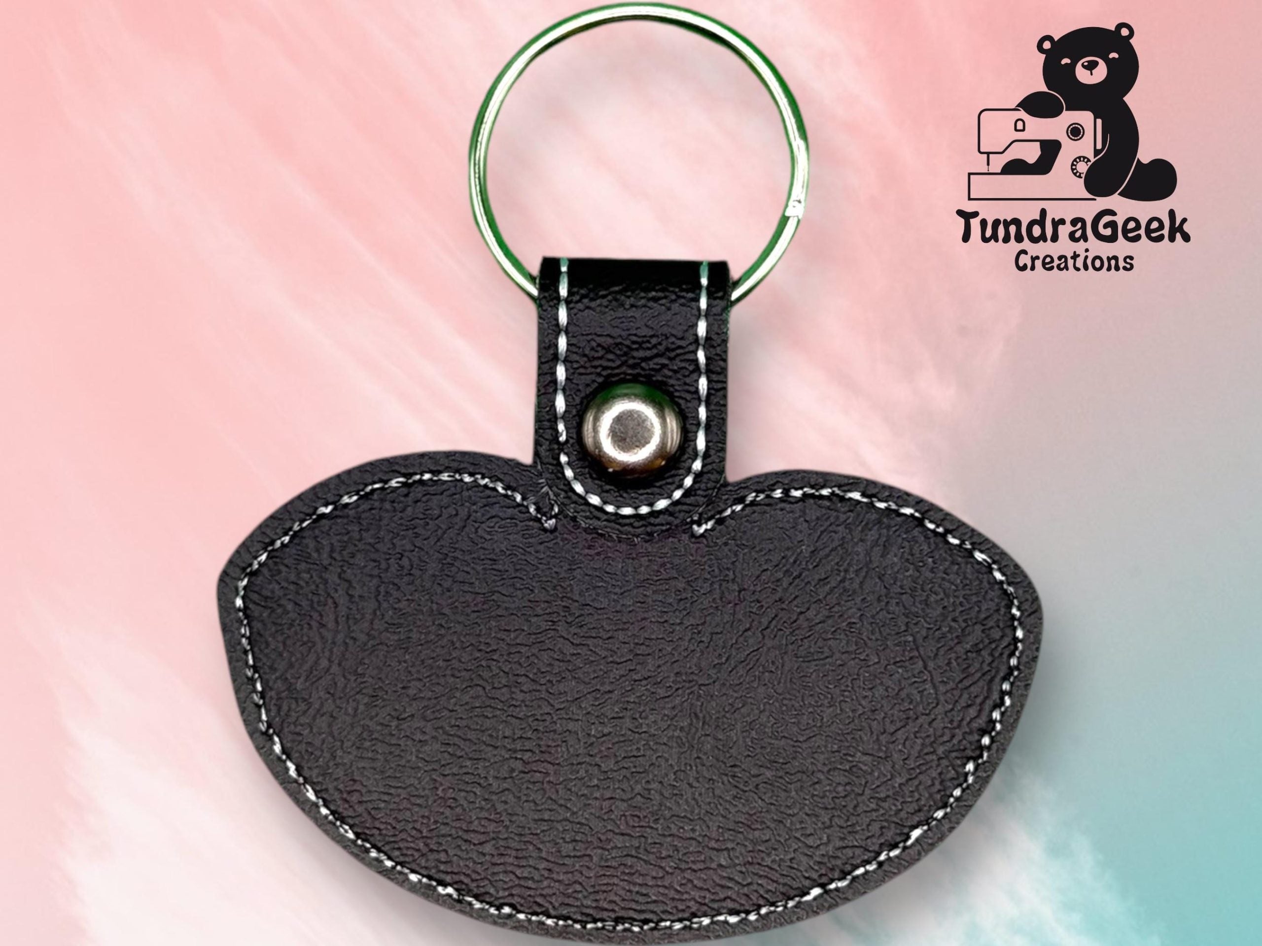 A black leather keychain with white stitching, shaped like a semicircle, is shown against a pink and blue background with the TundraGeek Creations logo in the top right corner.