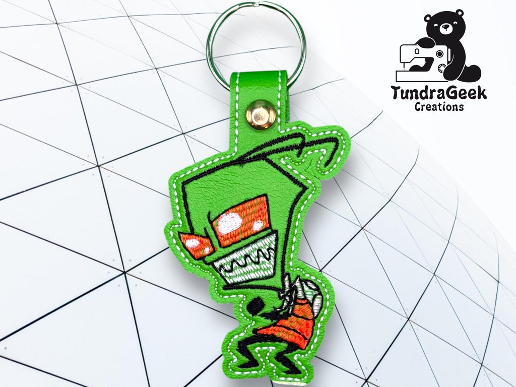 A green keychain featuring a cartoon alien character with red eyes and sharp teeth, placed on a geometric white background. TundraGeek Creations logo is visible in the corner.