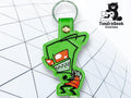 A green keychain featuring a cartoon alien character with red eyes and sharp teeth, placed on a geometric white background. TundraGeek Creations logo is visible in the corner.