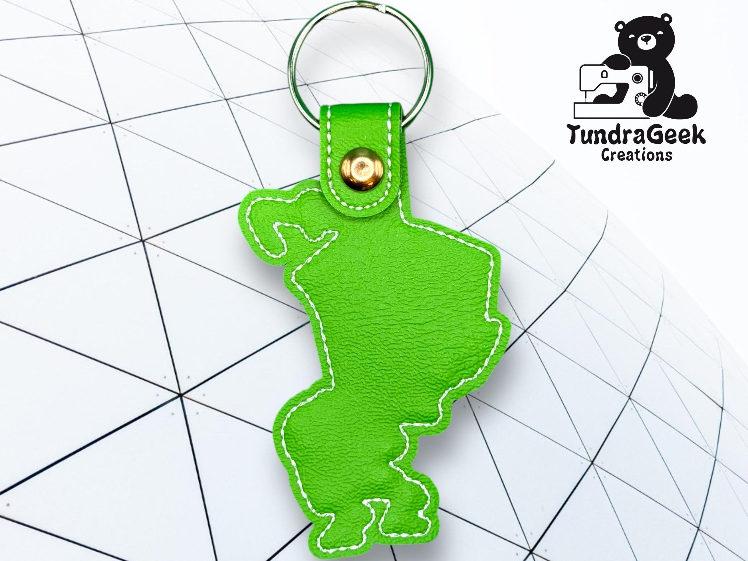 Green leather keychain in the shape of Michigans Upper Peninsula, with white stitching, on a geometric background. TundraGeek Creations logo is in the top right corner.