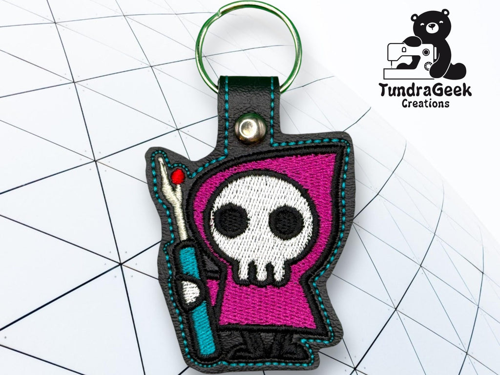 A keychain featuring a Grim Reaper cartoon character with a purple hood and a staff, displayed on a geometric patterned background. TundraGeek Creations logo is in the top right corner.