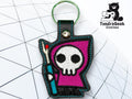 A keychain featuring a Grim Reaper cartoon character with a purple hood and a staff, displayed on a geometric patterned background. TundraGeek Creations logo is in the top right corner.