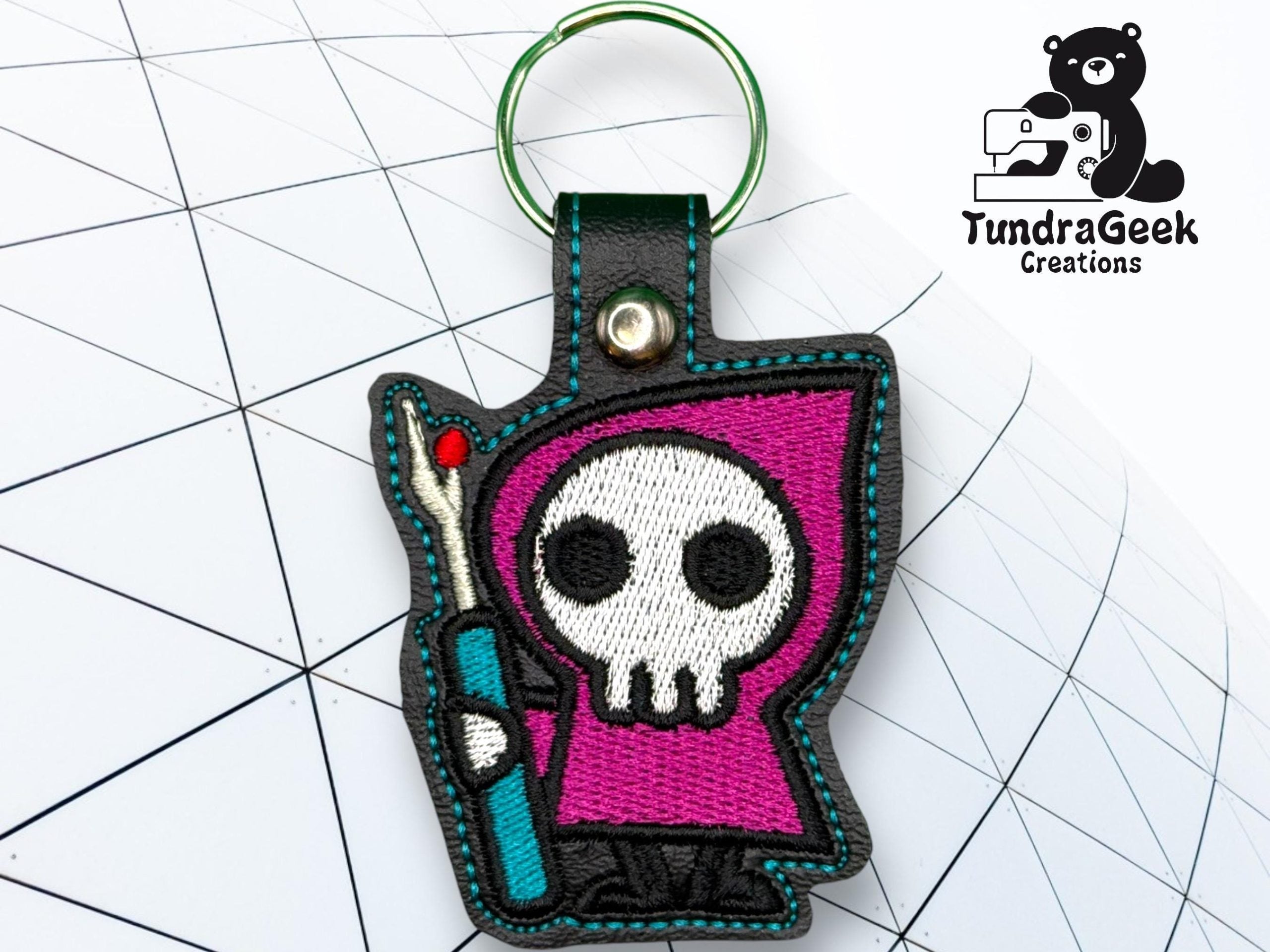 A keychain featuring a Grim Reaper cartoon character with a purple hood and a staff, displayed on a geometric patterned background. TundraGeek Creations logo is in the top right corner.