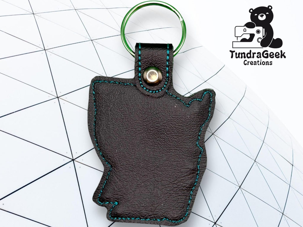 A leather keychain in the shape of a U.S. state with teal stitching and a green keyring is placed on a geometric background, next to the TundraGeek Creations logo.